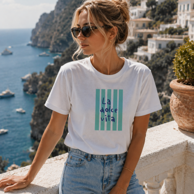 Mulher de t-shirt branca com texto La dolce vita e óculos escuros, junto a um corrimão com vista para mar e casas na encosta.