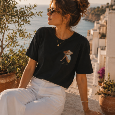 Mulher com t-shirt preta ilustrada e calças brancas sentada num muro exterior com plantas e paisagem de mar ao fundo