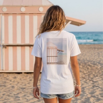 Pessoa vista de costas usando t-shirt branca com imagem de cabine de praia rosa e branca, praia ao fundo.