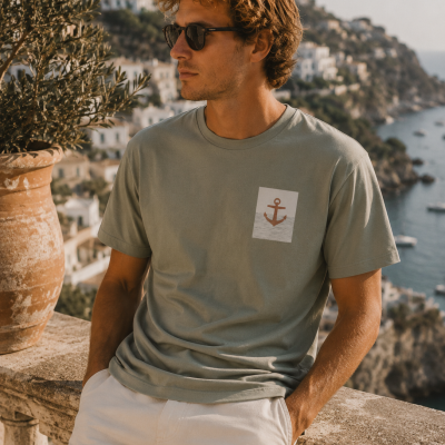 Homem com t-shirt verde-acinzentada com âncora, calças brancas e óculos, em varanda com vista para mar e casas.