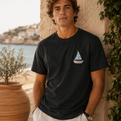Homem com t-shirt preta com bordado de veleiro, calças claras, junto a parede de pedra e vaso grande com planta, vista para o mar