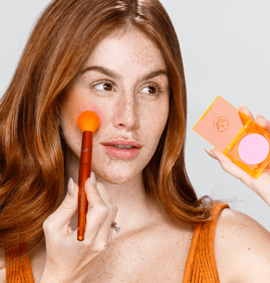 Pincel para Contorno e Iluminação - Mari Maria Makeup Mulher ruiva a aplicar blush cor-de-rosa com pincel laranja e a segurar embalagem transparente laranja