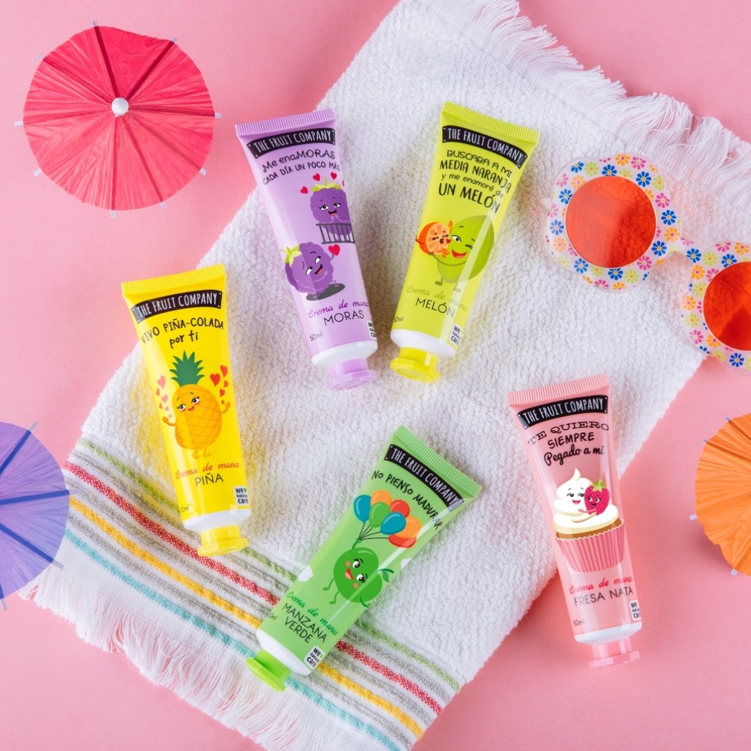 The Fruit Company - Creme de Mãos Cinco tubos coloridos da THE FRUIT COMPANY sobre toalha com guarda-sóis e óculos de sol em fundo rosa.