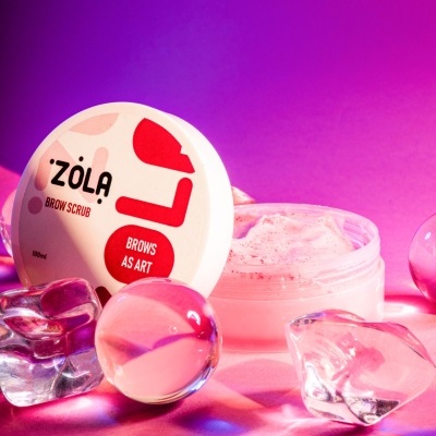 Frasco de creme de esfoliante para sobrancelhas rosa com tampa branca rotulada e objetos transparentes à volta