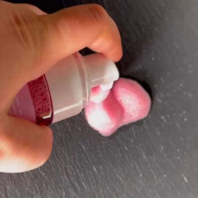 Mão a pressionar bico de embalagem branca a dispensar espuma rosa sobre superfície escura
