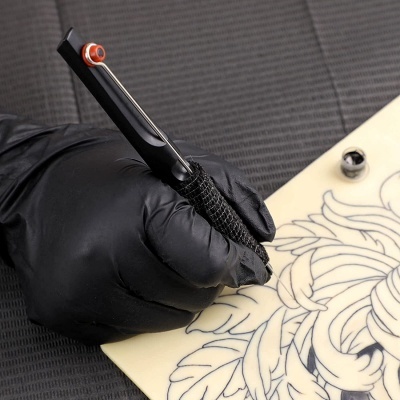 Mão com luva preta segurando máquina de tatuagem a trabalhar em pele sintética creme com desenho a preto.