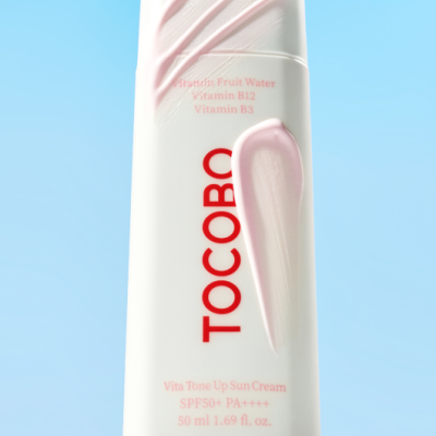 Frasco branco de creme solar TOCOBO com texto vermelho e rosa e creme rosa aplicado, fundo azul claro