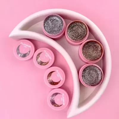 Cinco frascos de gel de brilho para unhas rosa com glitter prateado e dourado numa bandeja branca curva sobre fundo cor-de-rosa