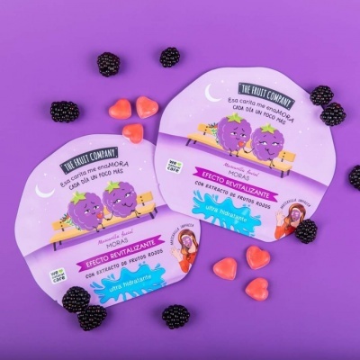 Pacotes lilases de máscara facial hidratante da THE FRUIT COMPANY com ilustração de amoras caricatas e corações cor de rosa sobre fundo roxo.