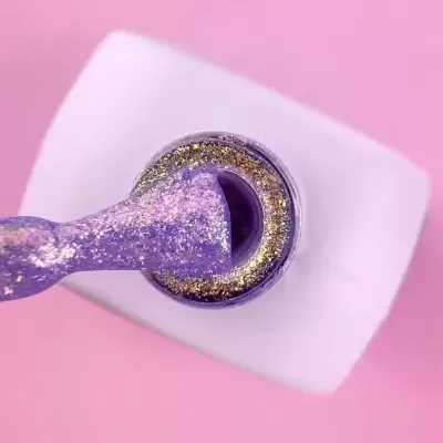 Verniz lilás com brilhos dourados num frasco branco aberto sobre fundo rosa.