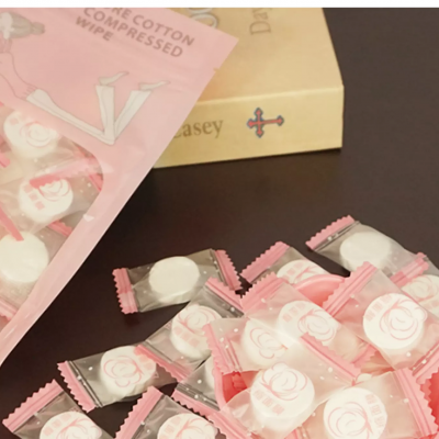 Pacote e muitas toalhitas de algodão comprimido embaladas individualmente com desenho de rosa em superfície escura e livro ao fundo.