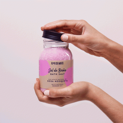 Frasco de sal de banho rosa com tampa preta e etiqueta com texto, segurado por mãos femininas