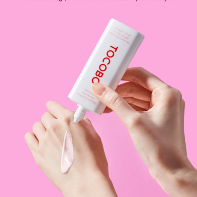 Bisnaga branca com creme rosa a ser aplicado na mão sobre fundo rosa