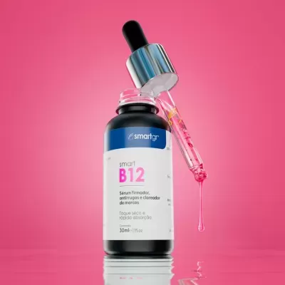 Frasco preto de sérum Smart B12 com conta-gotas a pingar líquido rosa, fundo rosa.