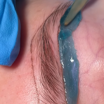 Aplicação de gel azul numa sobrancelha com espátula e mão com luva azul