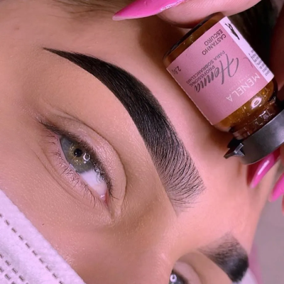 Frasco de óleo para sobrancelhas castanho escuro próximo a sobrancelha preta bem definida e mão com unhas rosa