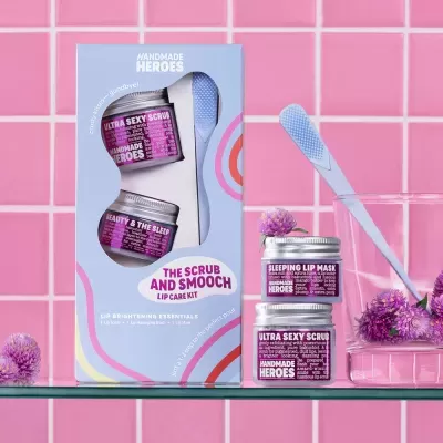 Kit de cuidado labial Handmade Heroes com frascos roxos e aplicador azul em fundo de azulejos rosa.