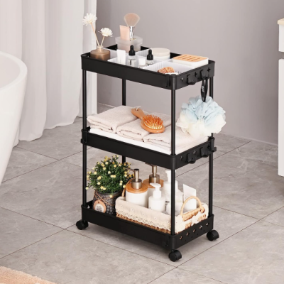 Carrinho preto com três prateleiras com toalhas, produtos de higiene, escovas e flores, contra fundo de chão cinza e cortina branca