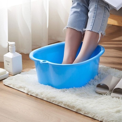 Bacia azul para banho de pés com pés dentro, tapete branco, frasco de sabonete e chinelos cinzentos