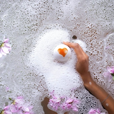 Mão segurando bola de banho branca com coração laranja em banheira com espuma e flores rosas