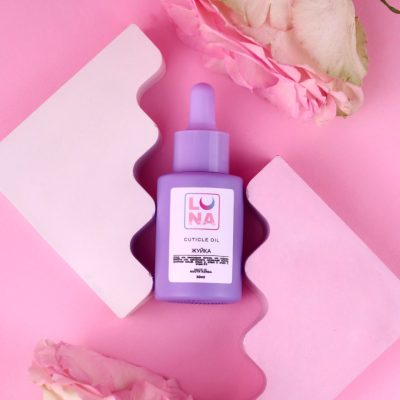 Frasco lilás de óleo para cutículas LUNA com conta-gotas sobre fundo rosa e decorado com flores