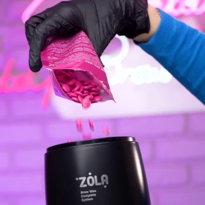 Máquina preta de aquecer cera ZÖLA com grãos rosados sendo despejados por pessoa com luvas pretas