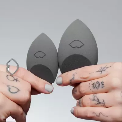 Duas esponjas de maquilhagem em forma de folha cinza com tatuagens nas mãos a segurá-las