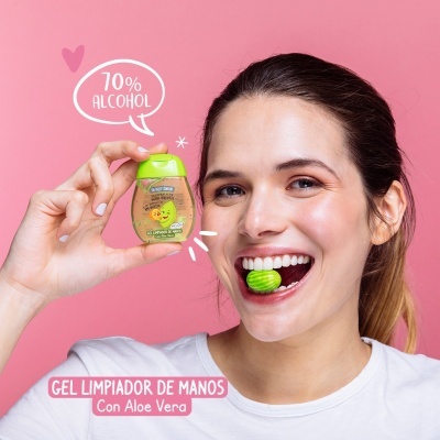 Mulher sorridente segura gel limpador de mãos com texto 70% ALCOHOL e doce verde na boca, fundo rosa.