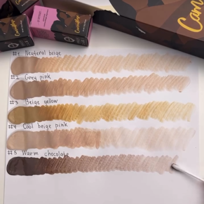 Cinco tons de corretor de maquilhagem com nomes escritos à mão e embalagens Castanhola na mesa