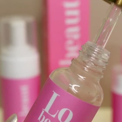 Frasco de gloss labial rosa com conta-gotas e tampa dourada num cenário com outros produtos semelhantes