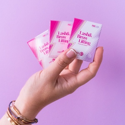 Caixas de Lash & Brow Lifting seguradas por uma mão com pulseiras sobre fundo lilás