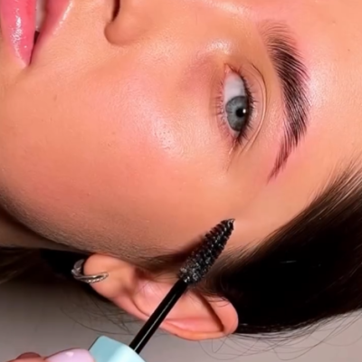 Pessoa aplicando máscara de pestanas preta nos cílios e sobrancelhas com fundo neutro