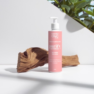 Frasco de mousse de limpeza facial rosa com texto branco em base branca, tronco de madeira e planta ao fundo