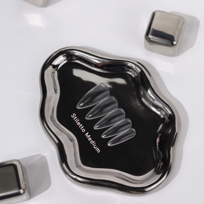 Unhas postiças transparentes stiletto medium sobre prato metálico prateado ondulado com pedras metálicas quadradas no fundo branco.