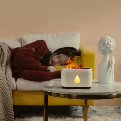 Mini lareira eletrónica branca sobre mesa ao lado de escultura, com mulher a dormir em sofá ao fundo