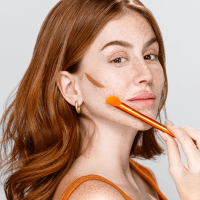 Mulher ruiva a aplicar maquilhagem cremosa castanha no rosto com pincel laranja