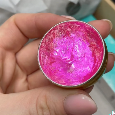 Recipiente metálico aberto com creme rosa brilhante segurado por mão com unhas pintadas de nude.