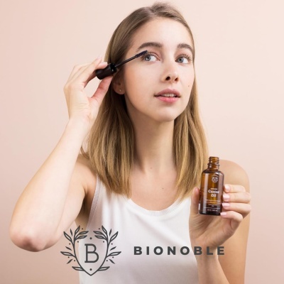 Mulher aplicando cosmético para sobrancelhas segurando frasco castanho com rótulo e logótipo BIONOBLE.