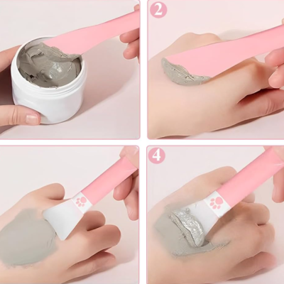 Envase de creme cinzento sendo retirado com espátula rosa e aplicado na mão com pincel rosa e decorado sob fundo claro