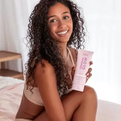 Mulher com lingerie bege segurando tubo rosa de produto GRADUAL GLOW numa cama