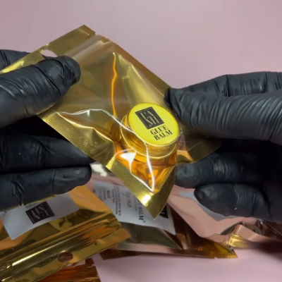 recipiente amarelo de GELÉE BALM em embalagem plástica dourada segurada por luvas pretas