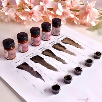 Frascos de esmaltes castanhos e pretos com amostras de cor e rótulos rosas sobre mesa branca com flores cor-de-rosa ao fundo