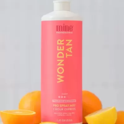 Frasco de spray profesional rosa com texto amarelo e branco, frutas cítricas à volta num fundo cinza claro.