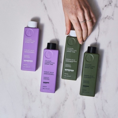 Frascos de spray correcção de cor para bronzeamento em bases violeta e verde sobre mármore claro, com mão a segurar um frasco verde.