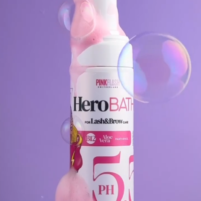 Frasco branco com espuma rosa e texto Hero BATH para Lash & Brow em fundo lilás com bolhas
