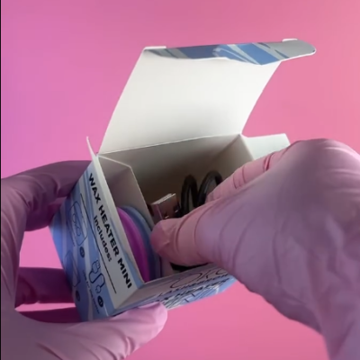 Mãos com luvas rosa segurando caixa aberta com aquecedor de cera mini