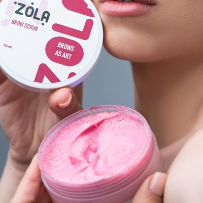 Recipiente de creme esfoliante para sobrancelhas cor-de-rosa aberto com tampa com texto da marca Zõla e lábios de pessoa em fundo cinza.