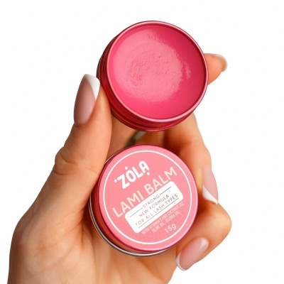 Creme labial rosa ZÔLA LAMI BALM numa embalagem redonda metálica segurada por uma mão.
