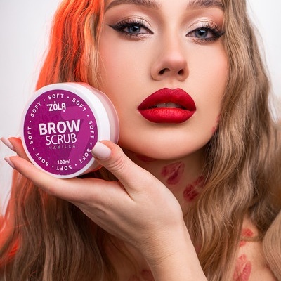 Produto Zola Brow Scrub Vanilla 100ml em embalagem rosa e branca segurado por mulher com batom vermelho.