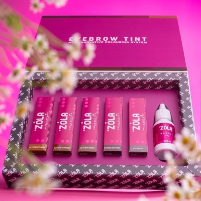 Conjunto de coloração para sobrancelhas Zola em embalagens rosa e branco sobre fundo rosa com flores brancas.